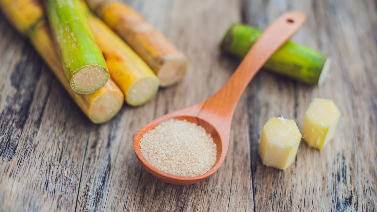 Sugar crystals in a wooden spoon beside cut sugarcane on a wooden surface